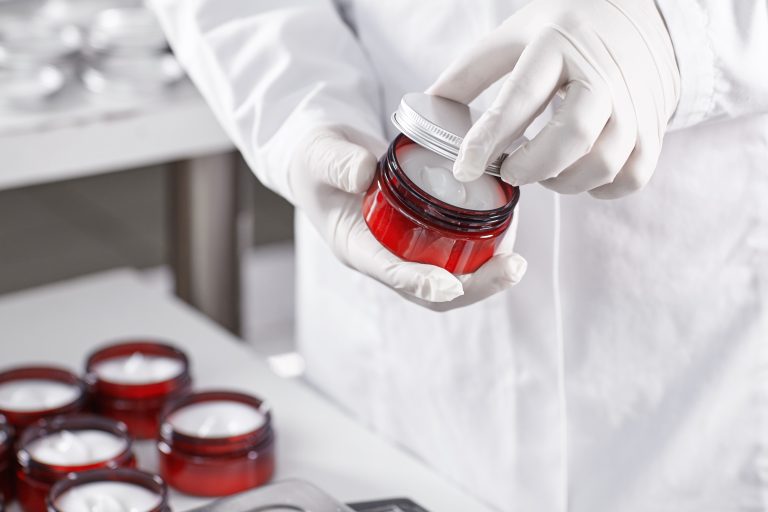 Factory worker in white gown and gloves holding red container with skin care product touching it with finger testing quality. Representing new product on factory